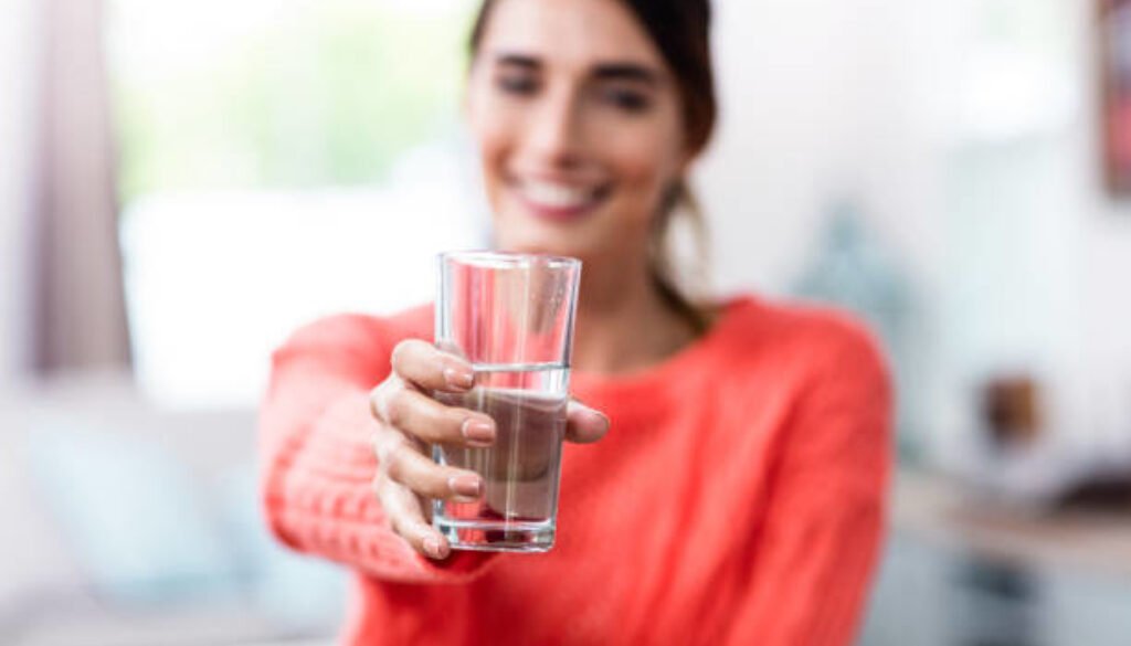 Happy young woman showing drinking glass with water at home